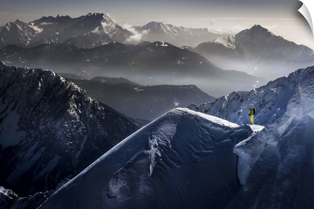 A skier pauses at the top of a mountain, with peaks of neighboring mountains in the background.