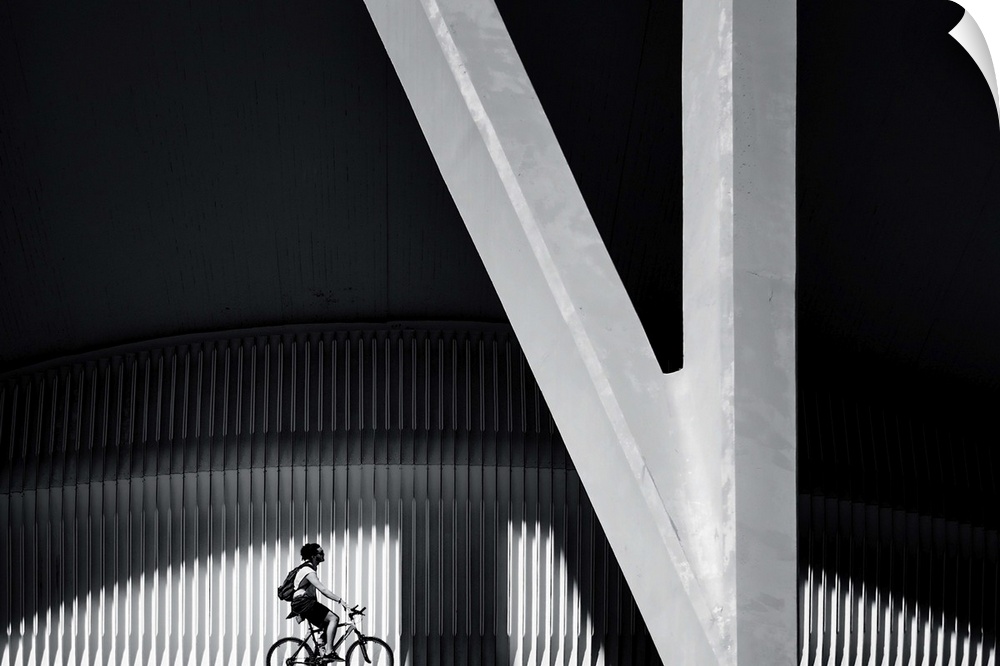 A person on a bicycle rides near a large concrete pillar and a ridged wall, forming an abstract image.