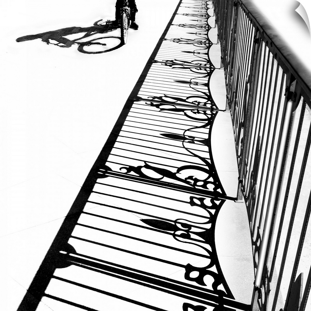 Black and white photograph of a bicycle and fence shadow onto the ground on a bright day.