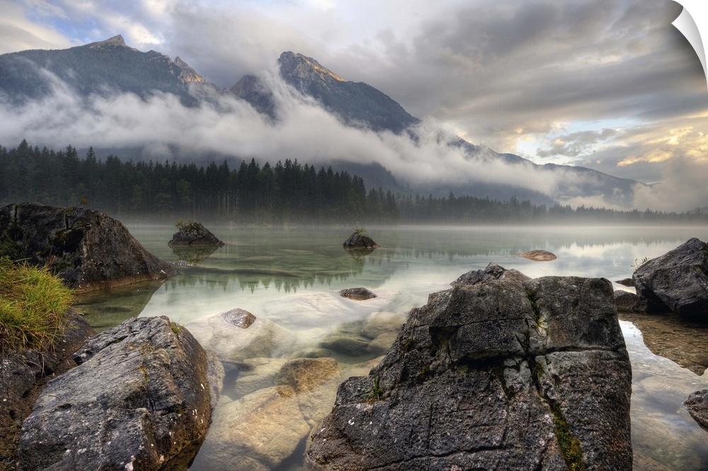 Rocks on the shore of a lake below mountains obscured by clouds, Germany.