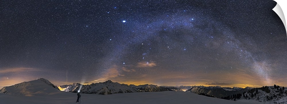 A panoramic photograph of a person standing in a desolate mountainous snowscape under a starry night sky, with the milky w...