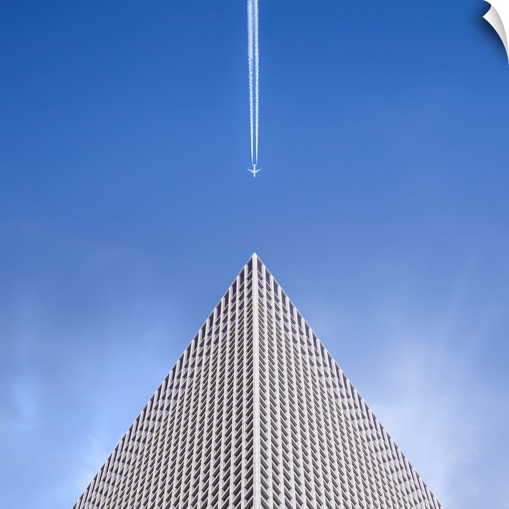 Looking up at the top of a skyscraper with an airplane leaving a vapor trail behind him.