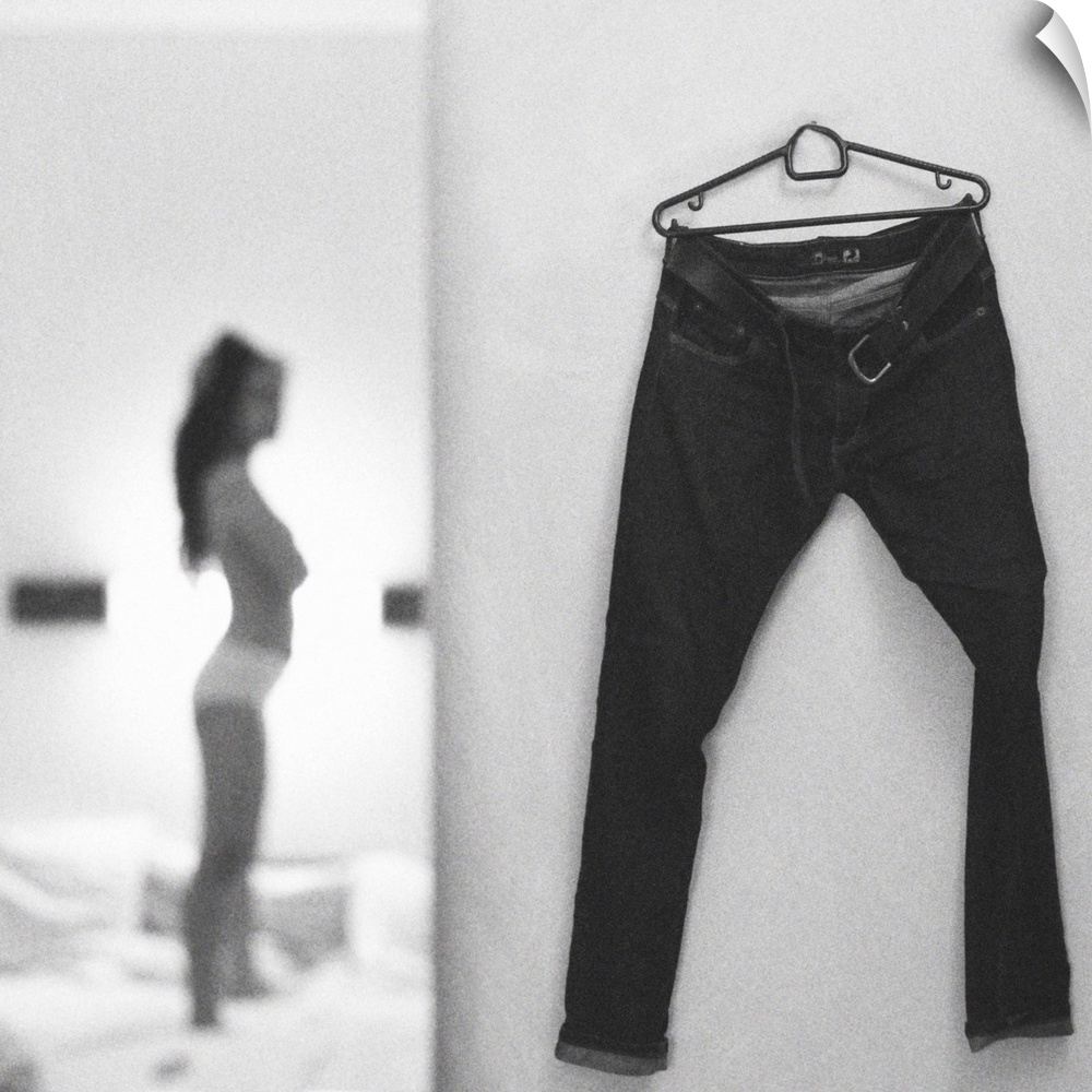 A focus on a pair of pants hanging on a wall while a young woman in the background gets ready to put them on.