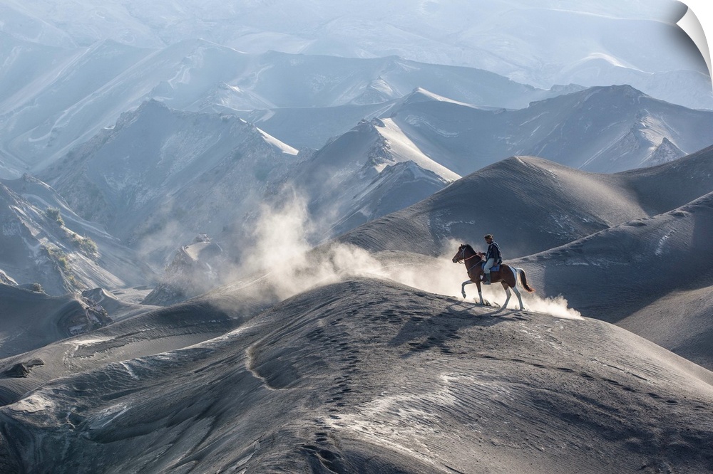 A man on a horse stands on the peak of a large hill in a valley surrounded by dunes.
