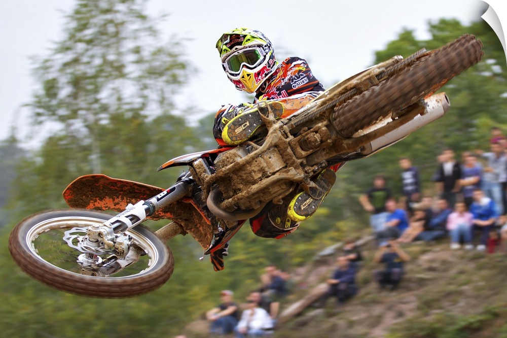 Motocross biker Tony Cairoli jumping his bike as a crowd watches in the background, Maggiora, Italy.
