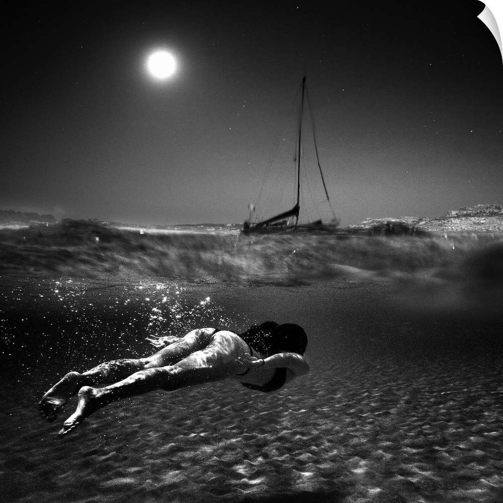Black and white photo of a woman in a bikini swimming just underneath the surface of the water near a boat, under a full m...
