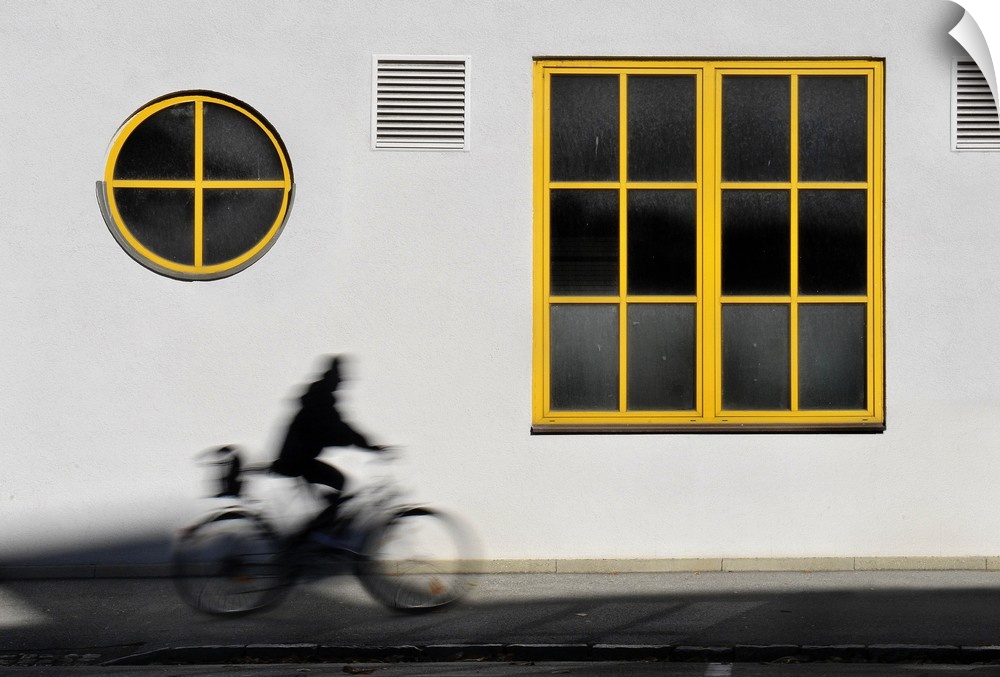 A person rides by a white wall with yellow trim windows on bicycle.