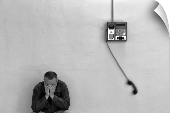 A man with his head in his hands leaning against a wall with a payphone swinging from the receiver.