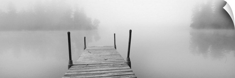 A black and white photograph of a dock stretching out over a foggy lake in an idyllic setting.