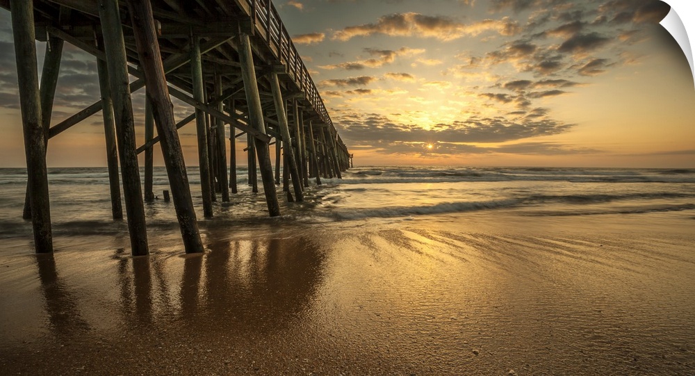 A photograph of a long pier jetting out over the ocean. Sunlight bathing the sky in an orange glow and the sun sets.