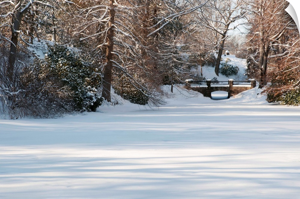 A snow-covered landscape with pond, trees; and a bridge.