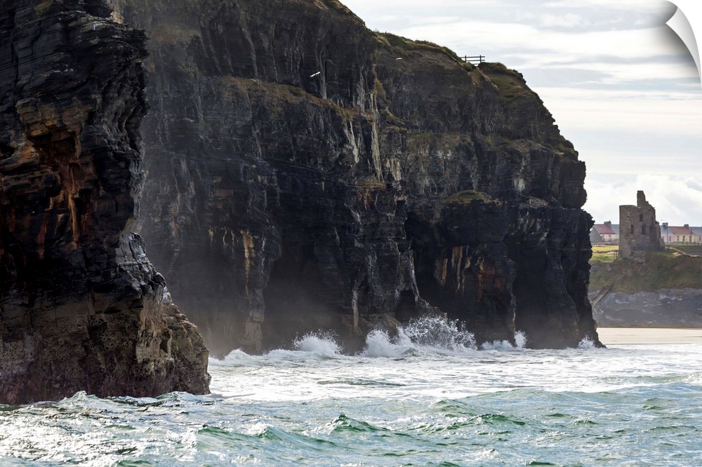 Dark straight cliffs with waves crashing into the rock with ruined castle turret in background, Ballybunion, County Kerry,...