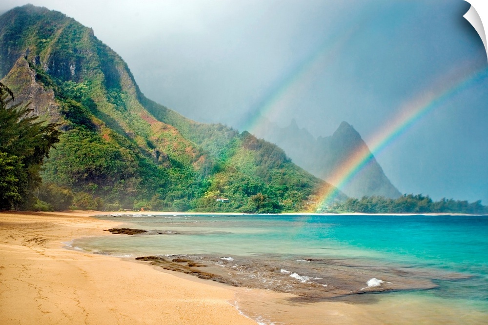 A landscape photograph with double rainbows on a tropical beach with mountains in the background.
