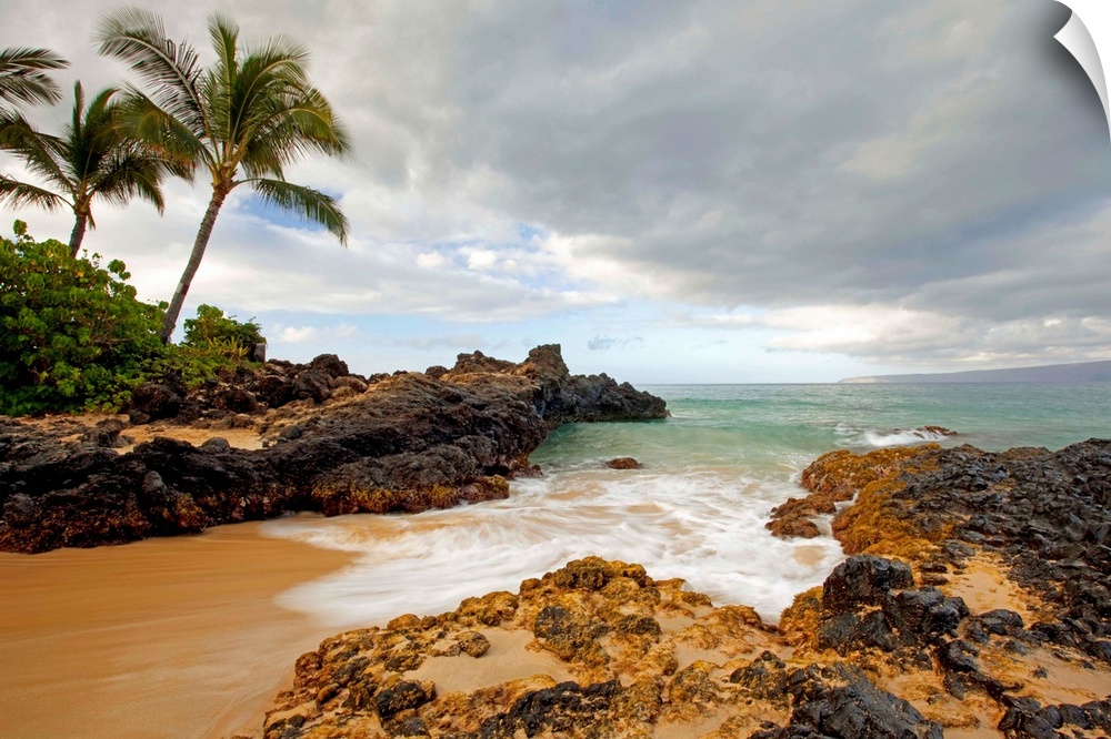 Photograph of a sandy break in the rocky coast as water rushes in to the cove with palm trees on an overcast day.