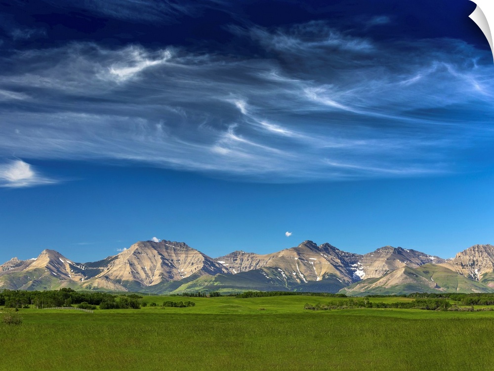 Mountain range with wispy white clouds, blue sky and green field in the foreground
