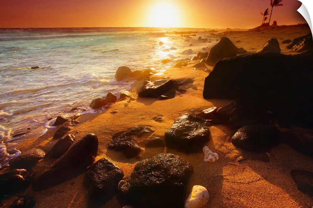Photograph taken of a sunset on the ocean horizon with various rocks spread out on the beach in the foreground.