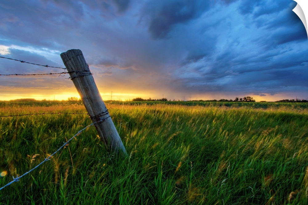 Photograph of wooden fence with barbwire in a field under dark storm clouds.