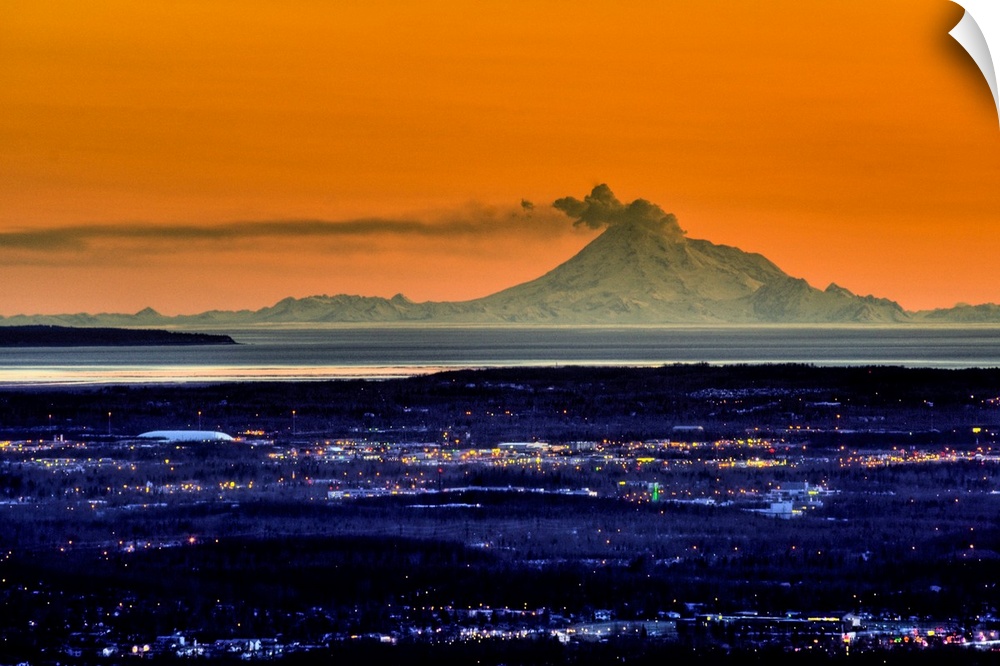 Ash billows out of an active volcano which looms over a city in the evening in the Aleutian mountain range.