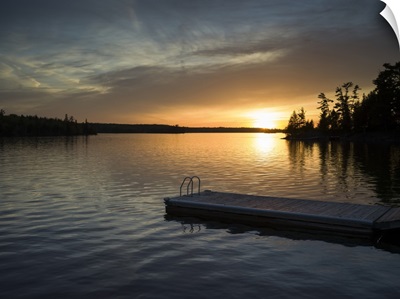 The sun setting over a tranquil lake and silhouetted trees with a dock in the foreground
