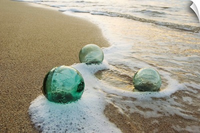 Three Glass Fishing Floats Roll On The Sandy Shoreline With Ripples Of Water And Seafoam