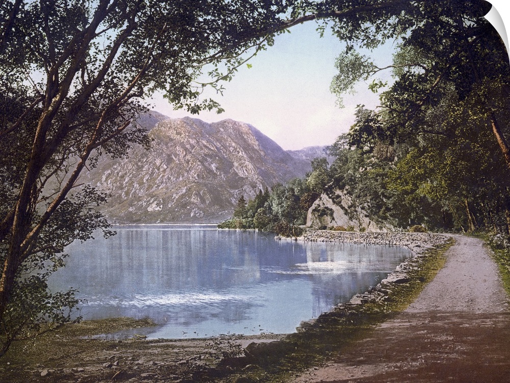A vintage shot of a gravel path winding its way at the water's edge as leafy trees and cliffs loom overhead.