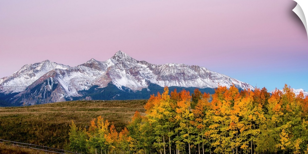 Landscape photograph of a snowy mountain range with colorful Fall trees in the foreground and a cotton candy sky above.