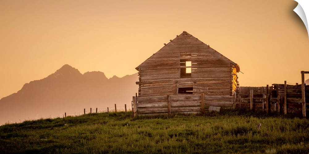 Warm photograph of an old wooden barn on a hilltop with a silhouette of mountains in the background.