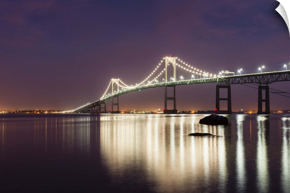 A photograph of a large suspension bridge seen lit up at night casting long reflections on the water below.