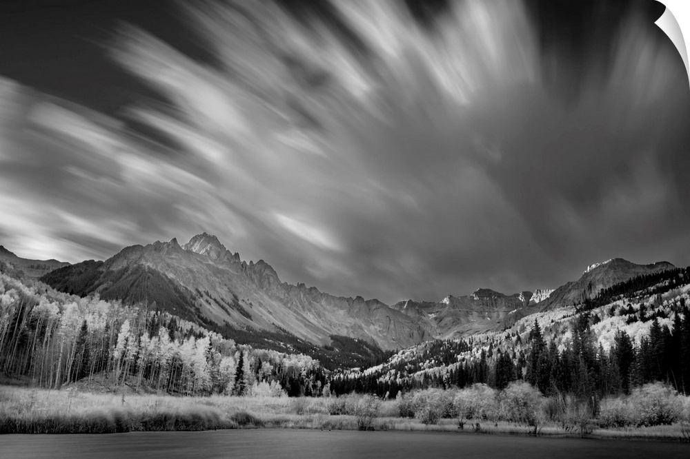 Black and white landscape photograph of a lake in front of a mountain range with blurred moving clouds in the sky.