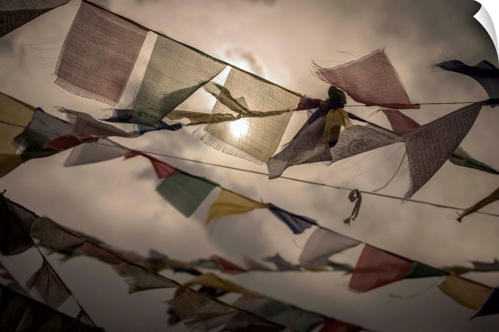 Photograph of colorful, worn prayer flags blowing in the wing with the sun shining through the clouds in the background.