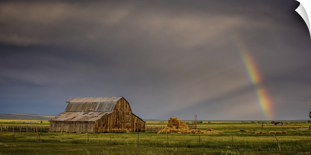 A photograph of an old barn in a wilderness landscape with a rainbow peaking out from clouds in the distance.