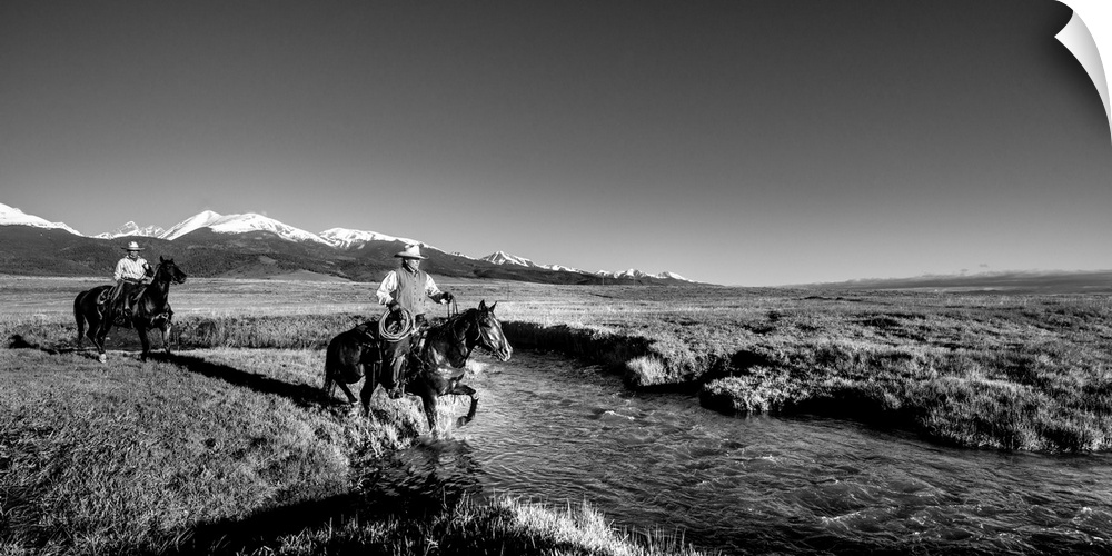 Black and white photograph of two cowgirls crossing a river on horseback with snow capped mountains in the distance.
