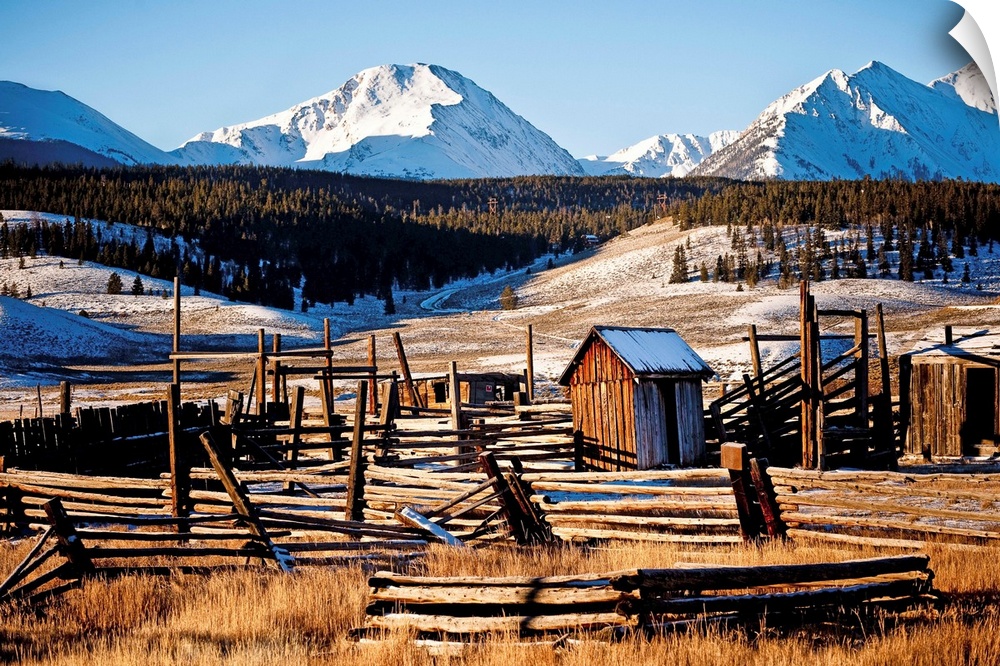 Landscape photograph of run down wooden structures in the valley surrounded by snow covered mountains.