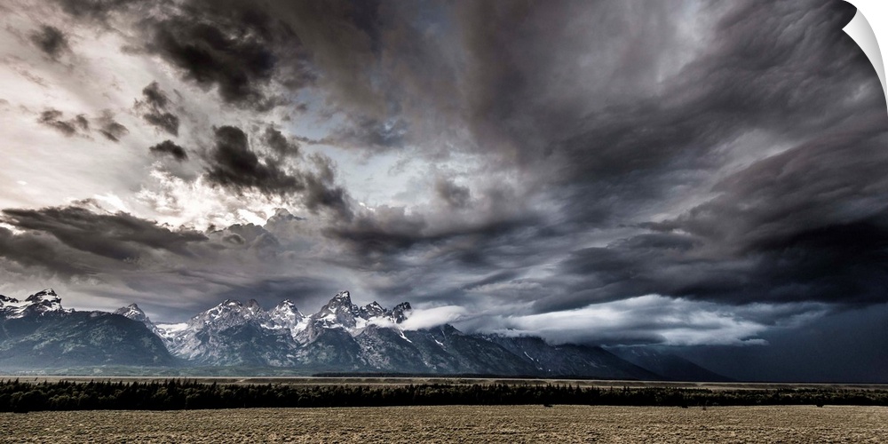 Landscape photograph of a field in front of snow capped mountains with a dramatic stormy sky above.