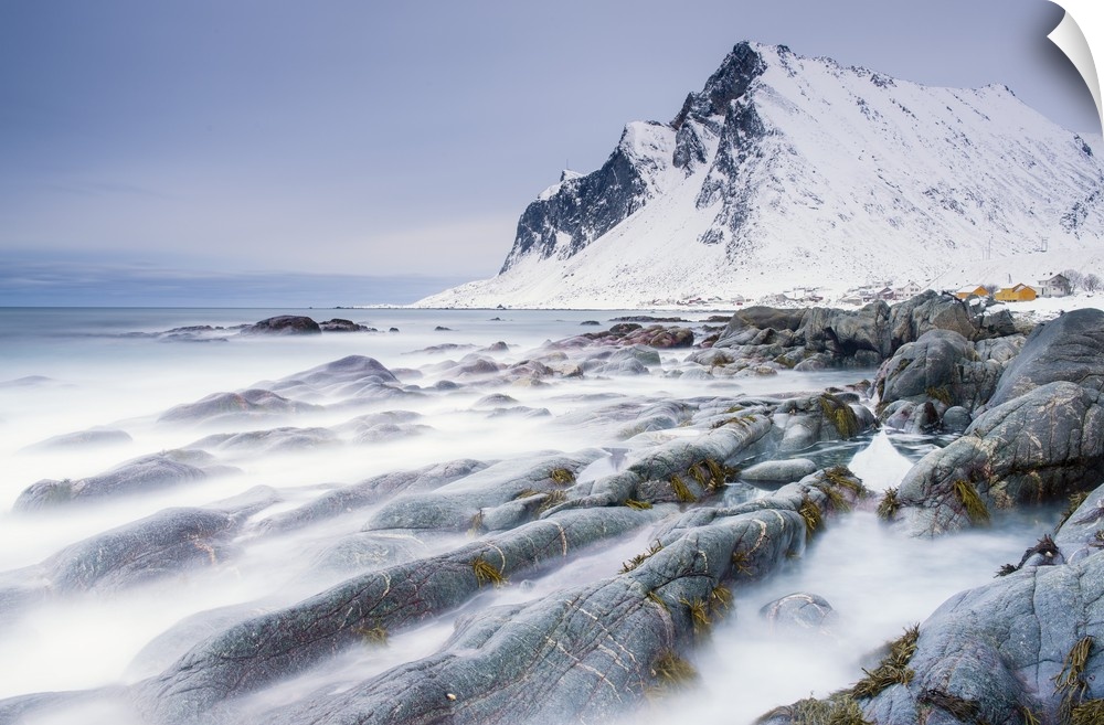 A photograph of a snow covered mountain seen from a rocky shoreline with motion blurred water caught in a long exposure.