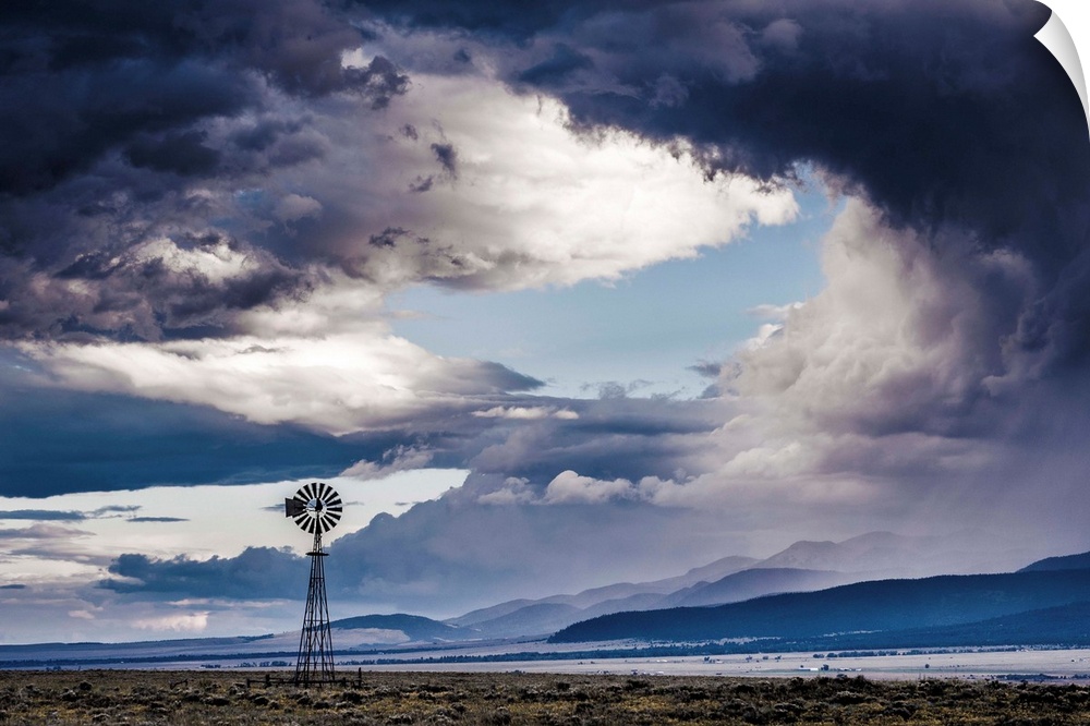 Cool toned landscape photograph of a windmill with a mountain range in the background and a dramatic sky.
