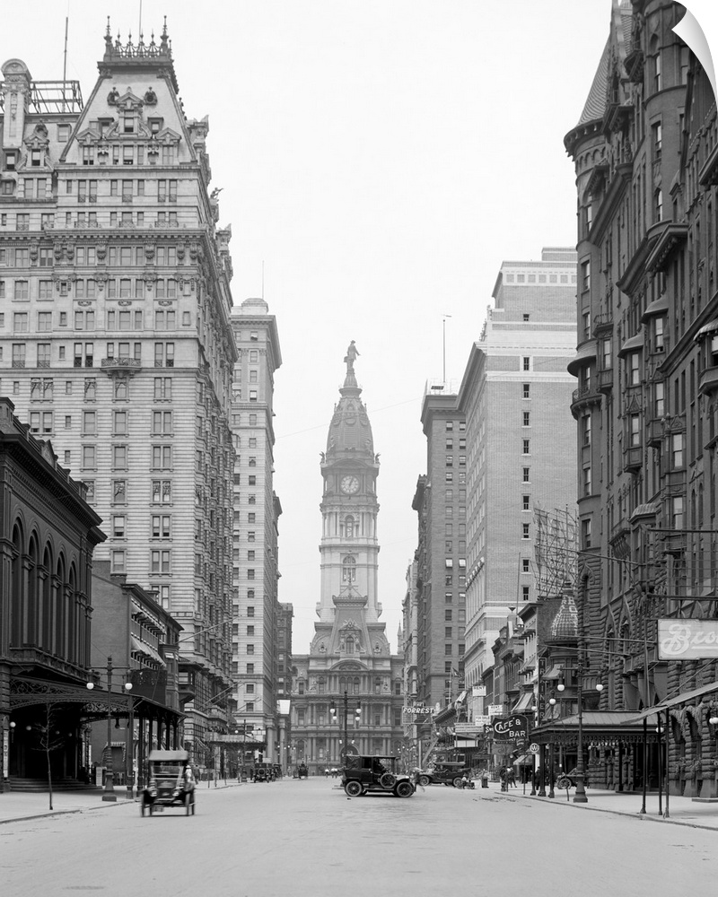 This is a vintage photograph in black and white of downtown Philadelphia. Tall buildings line the street that has cars dri...