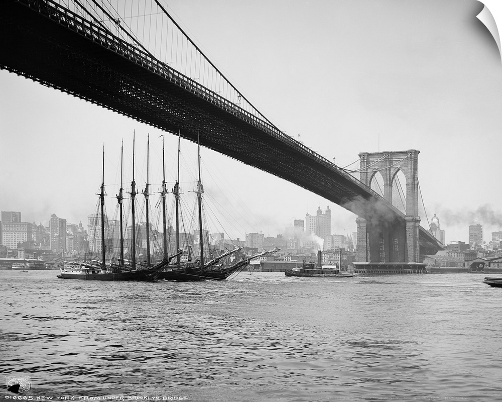 Horizontal, vintage photograph of an old ship passing beneath the Brooklyn Bridge, along the East River, the New York City...