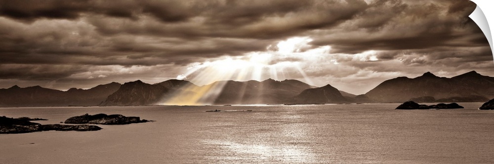 Panoramic photo, in a sepia tone, of the sun streaming through a heavy clouded sky over a body of water with mountains in ...