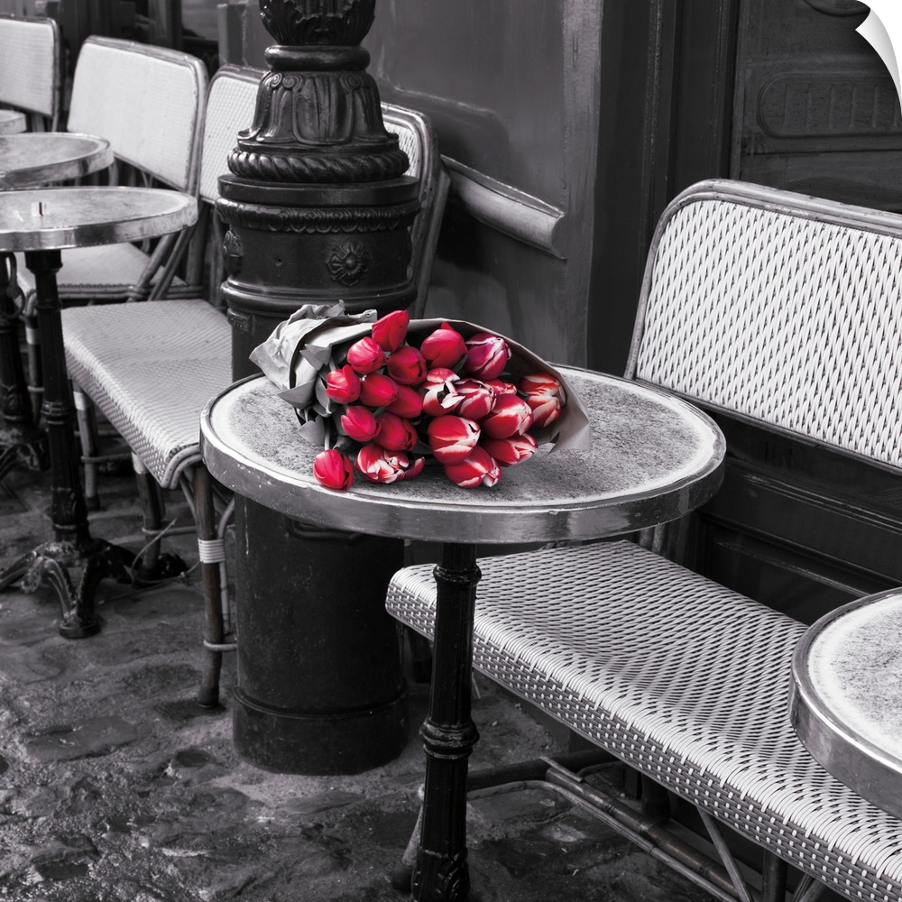 Square black and white photograph of sidewalk seating at a cafe with colored flowers on a table.