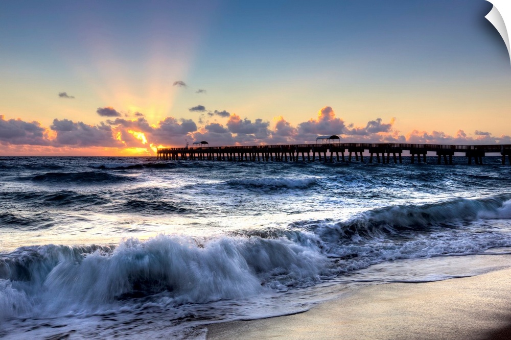A horizontal photograph of crashing waves on a beach with a sunset and pier in the background.