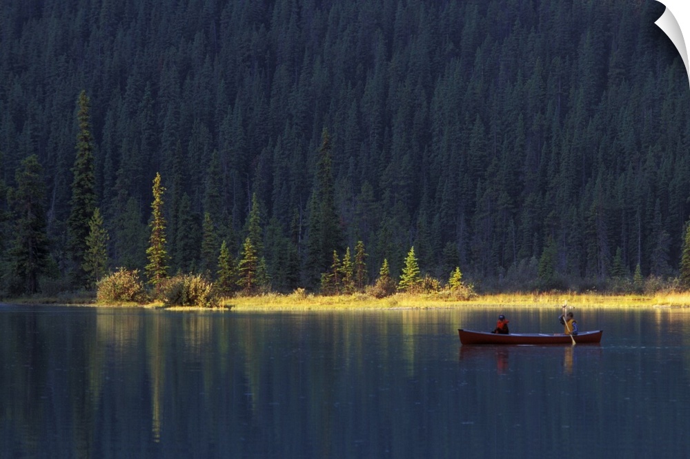 Two people paddle in a canoe in the early morning light, with lush forest behind them, on Waterfowl Lake, in Banff Nationa...