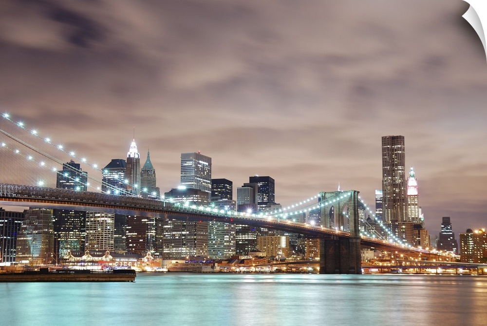 Panoramic view of Manhattan with Brooklyn Bridge at night and skyscrapers illuminated over Hudson River.