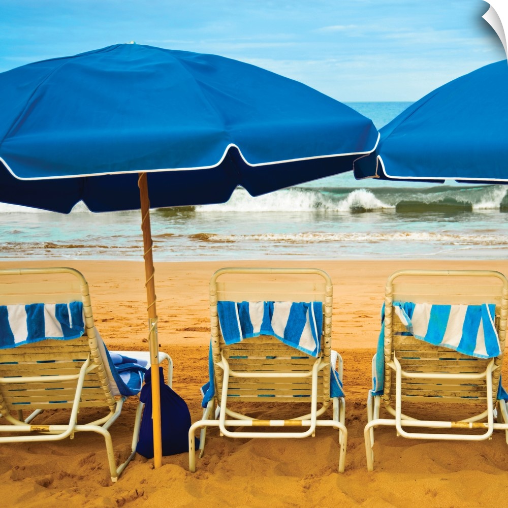 Square photograph of three beach chairs with blue umbrellas lined up right in front of the ocean.