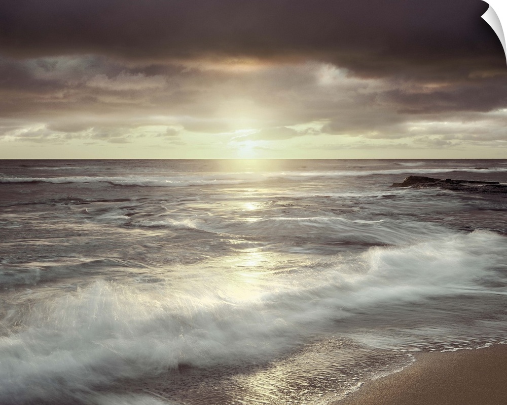 Long exposure photograph of waves crashing on the shore with an evening sunset and a cloudy sky.