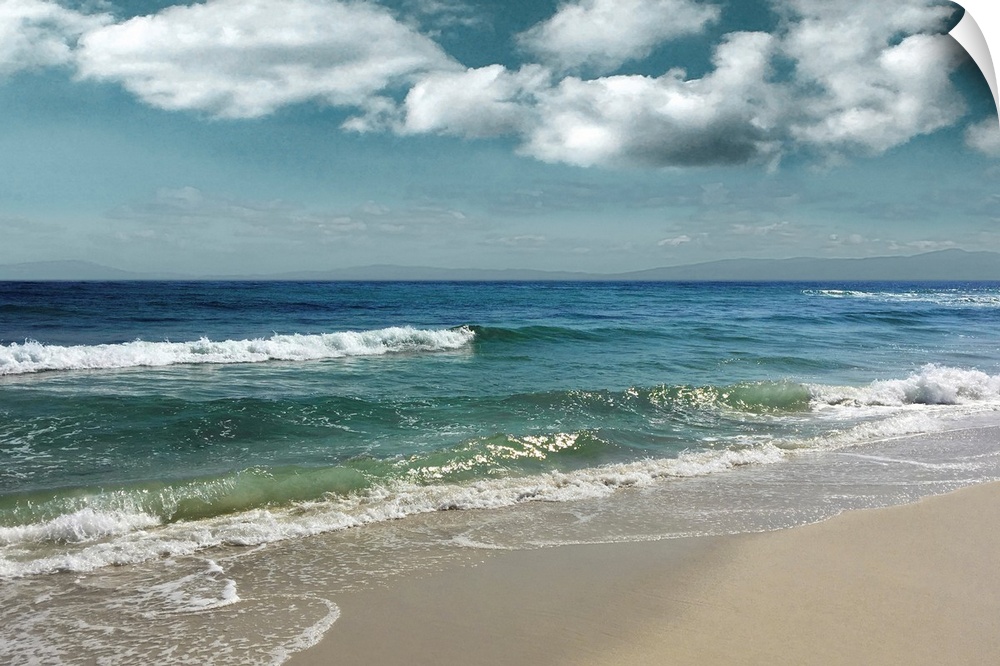 This serene photo shows rippling waves as they approach the beach with puffy white clouds in the background.