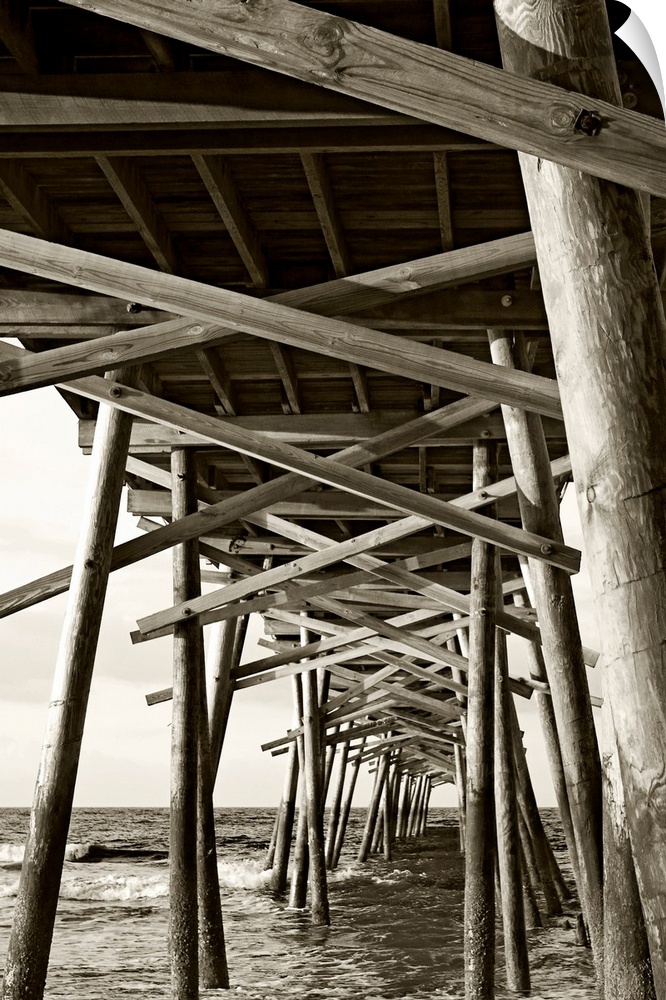 Big monochromatic photograph taken from beneath a large wooden pier examines the foundation of it while the ocean crashes ...