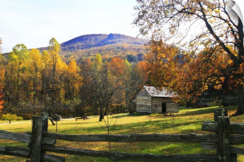 This is a landscape photograph of a log cabin in a meadow surrounded by autumn trees behind a primitive wood fence.