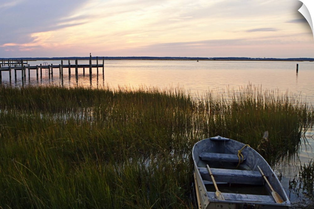A boat sits in shallow water amidst tall grass at dusk by a pier as calm waters reflect the sun's rays.