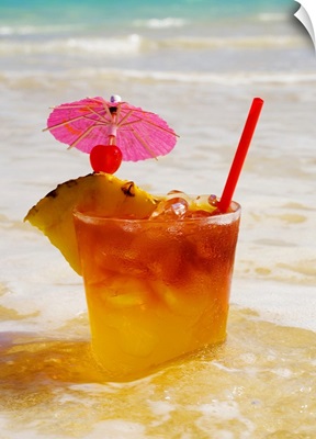 A mai tai garnished with pinapple and a cherry, sitting in shallow water on the beach.